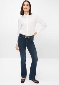 Chemise blanche à col, jean bleu foncé évasé, ceinture noire avec boucle en argent, et ballerines noires en mesh. Texture lisse dans l'ensemble.