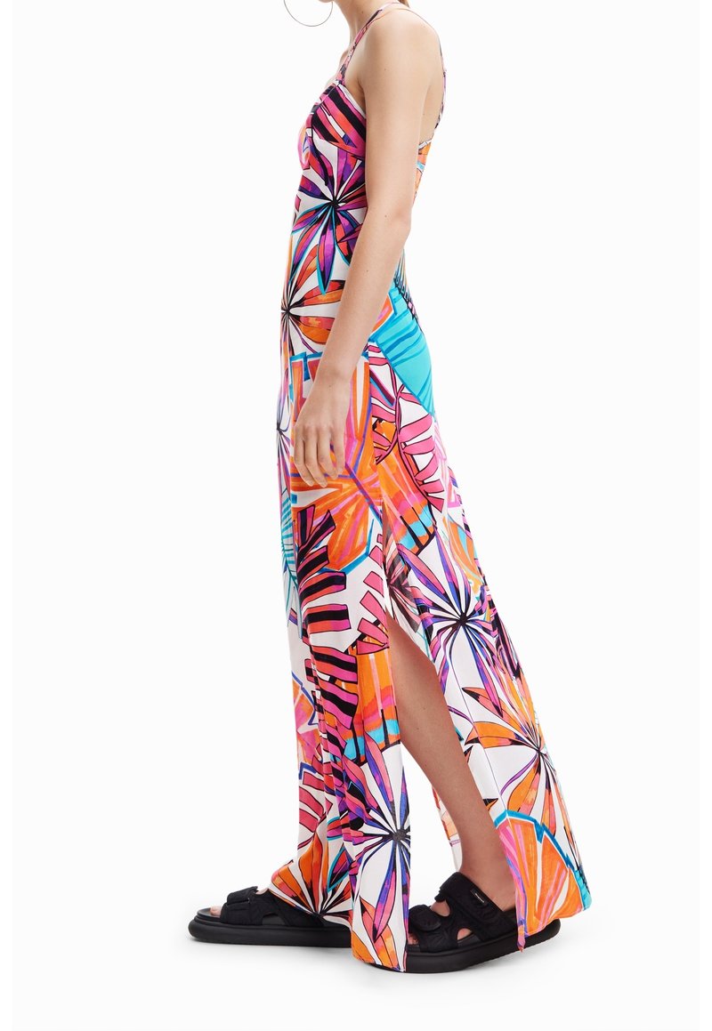 Femme portant une robe longue fleurie, colorée, sans manches, avec une fente latérale, associée à des sandales noires, debout de profil.