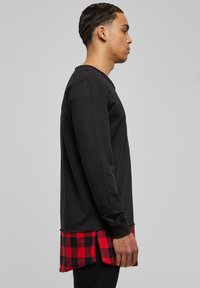 Chemise noire à manches longues avec une couche inférieure à carreaux rouges et noirs, présentant un ourlet incurvé et un col texturé.