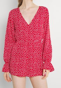 Femme portant une combinaison rouge à manches longues avec un motif floral blanc et un décolleté en V profond, montrée du cou au milieu des cuisses sur un fond uni.