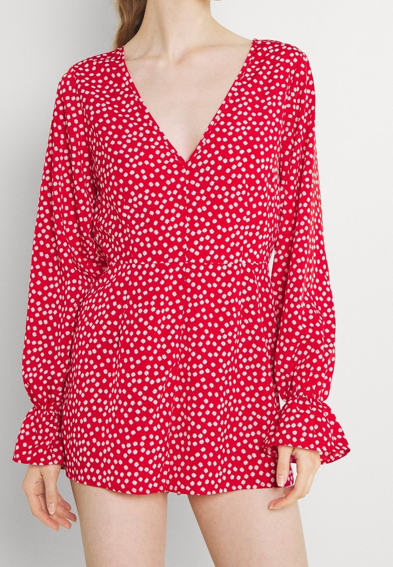 Femme portant une combinaison rouge à manches longues avec un motif floral blanc et un décolleté en V profond, montrée du cou au milieu des cuisses sur un fond uni.