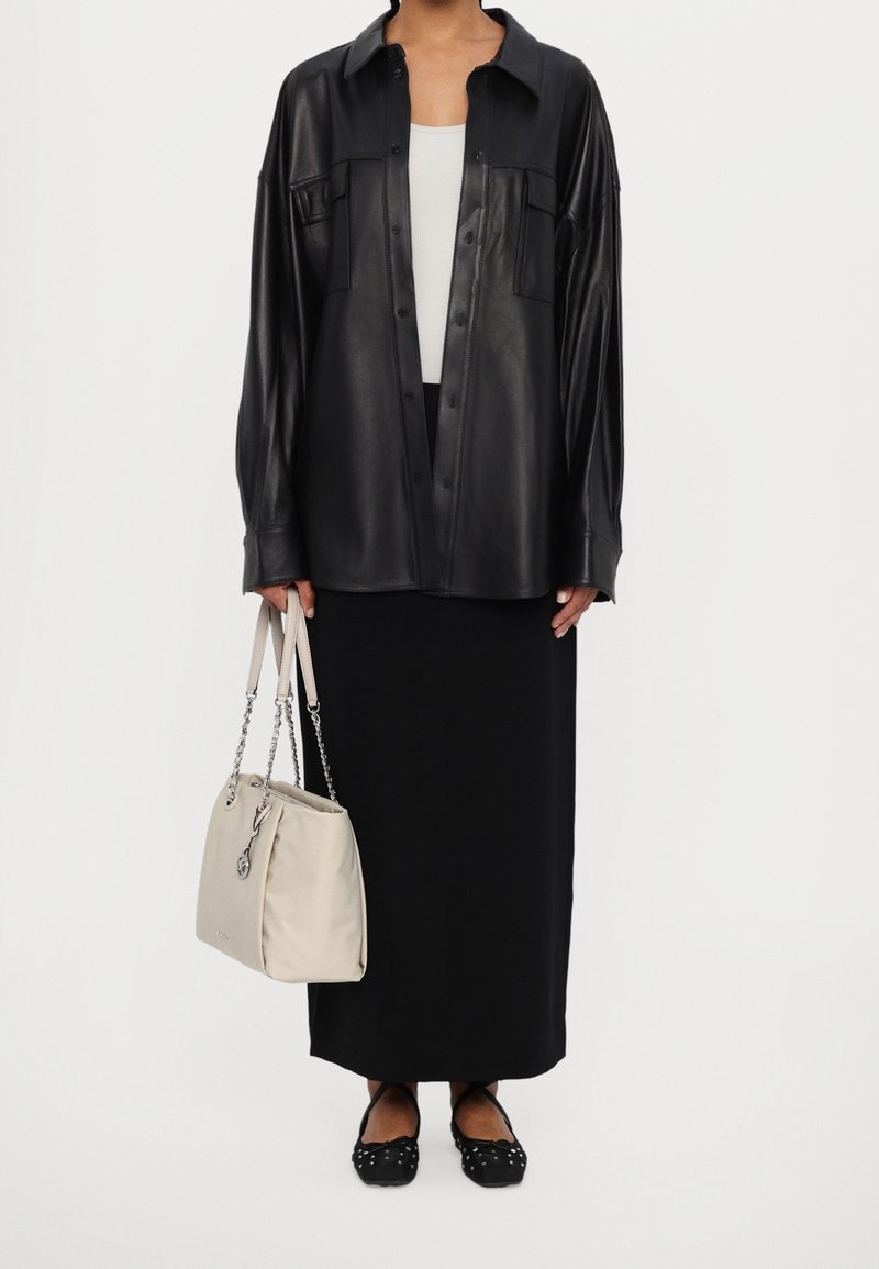 Personne portant une chemise en cuir noire à boutons, une longue jupe noire, des ballerines noires cloutées, et tenant un sac à main beige avec des anses en chaîne.