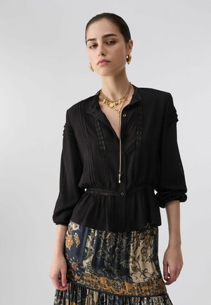 Femme aux cheveux plaqués en arrière, portant un chemisier noir, des colliers en or superposés, des boucles d'oreilles en or et une jupe à motifs floraux et abstraits.