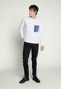 Bílá košile s dlouhým rukávem a modrou kapsou se zipem, v kombinaci s černými slim-fit kalhotami a černými botami. Jednoduchý design a barvy.