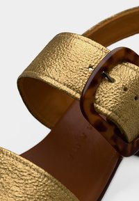 Κοντινή λήψη χρυσής υφής λουριού σανδάλου με ορθογώνια αγκράφα από όστρακο χελώνας σε καφέ βάση ποδιού.