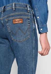 Jean en denim bleu foncé avec un patch en cuir "Wrangler" sur la poche arrière, avec des coutures distinctives et un design à coupe standard.