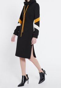 Svart klänning med cowl-krage, långa ärmar med orange och vita ränder, sidofraktioner och detaljer med dragsko. Bärs med svarta högklackade stövlar.