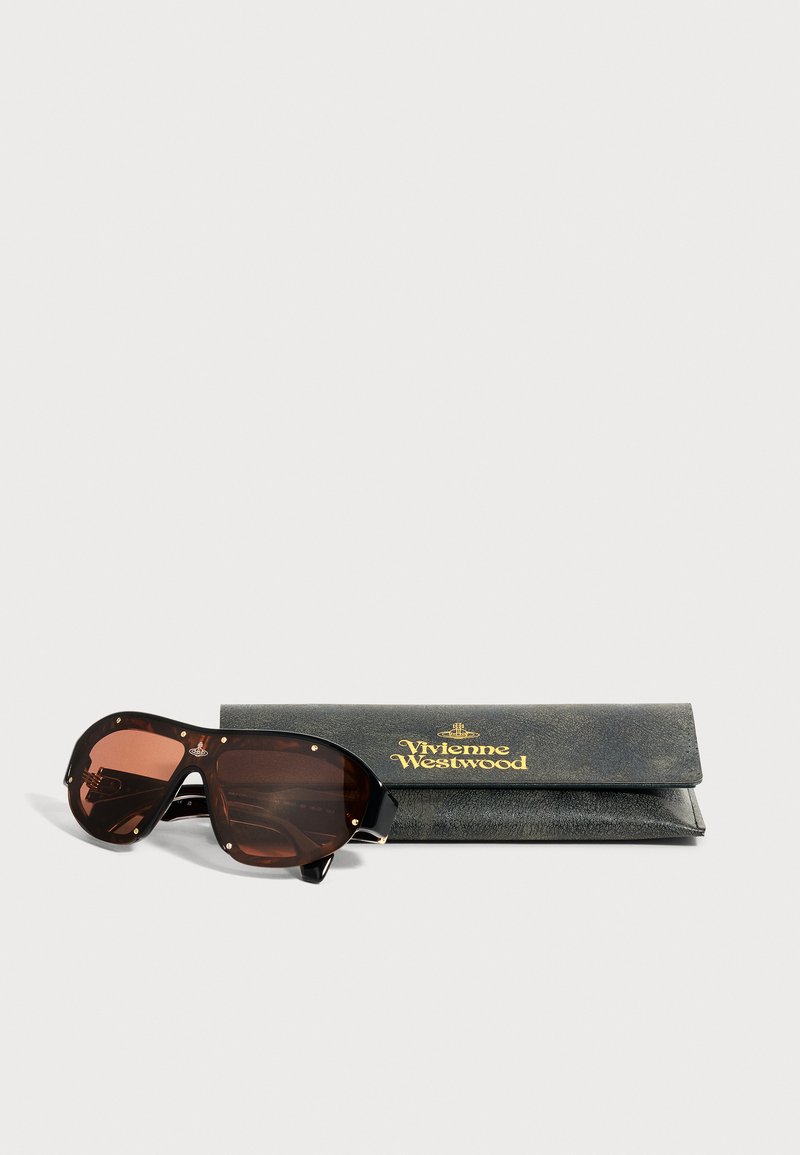 Occhiali da sole oversize marrone scuro con borchie dorate accanto a una custodia nera Vivienne Westwood con logo dorato.