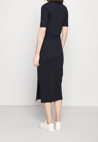 Marineblaues, knielanges Kleid aus weichem Stoff, mit kurzen Ärmeln, einem Gürtel in der Taille und einem seitlichen Schlitz. Getragen mit weißen Sneakers.