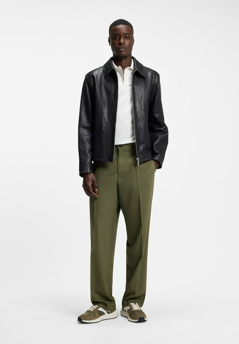 Homme portant une veste en cuir noire, un polo blanc, un pantalon vert olive et des baskets vertes et blanches, debout devant un fond blanc.