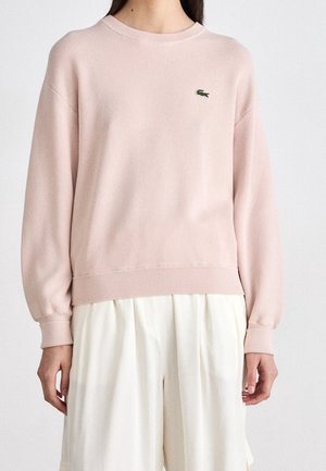 Femme portant un pull à manches longues rose clair avec un petit logo vert sur la poitrine gauche et un pantalon blanc plissé ample.