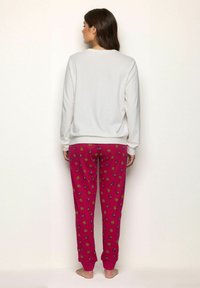 Mujer de pie, descalza, mirando hacia una pared, vestida con una camiseta de manga larga blanca y pantalones de pijama rojos con patrones amarillos y negros.