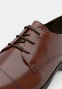 Chaussure habillée en cuir marron avec une finition brillante, dotée d'un système de laçage fermé, de détails cousus et d'une forme de bout arrondi.
