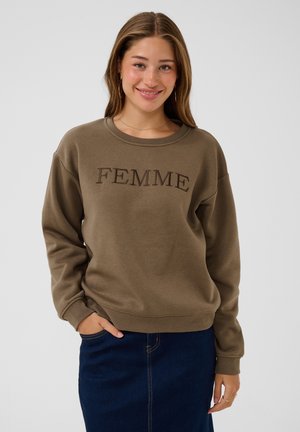 Jonge vrouw die lacht, draagt een bruine trui met de tekst "FEMME" en een donkerblauwe rok, staand tegen een effen lichte achtergrond.