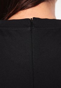 Schwarzes Oberteil mit Reißverschluss auf der Rückseite und kleinem Ziehgriff, aus einem glatten, dehnbaren Stoff gefertigt. Klare Linien mit minimalistischen Designelementen.