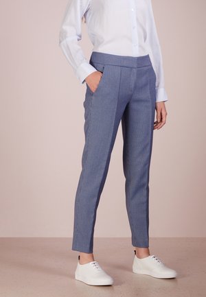 Vrouw draagt blauw geruite, taps toelopende broek, wit overhemd met knopen, witte sneakers, staand met één hand in haar zak.