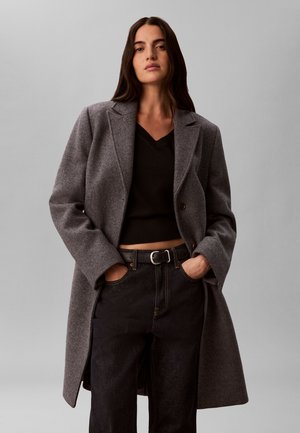 Femme aux longs cheveux foncés porte un manteau en laine gris, un pull court noir et un jean noir, les mains dans les poches, sur un fond gris uni.