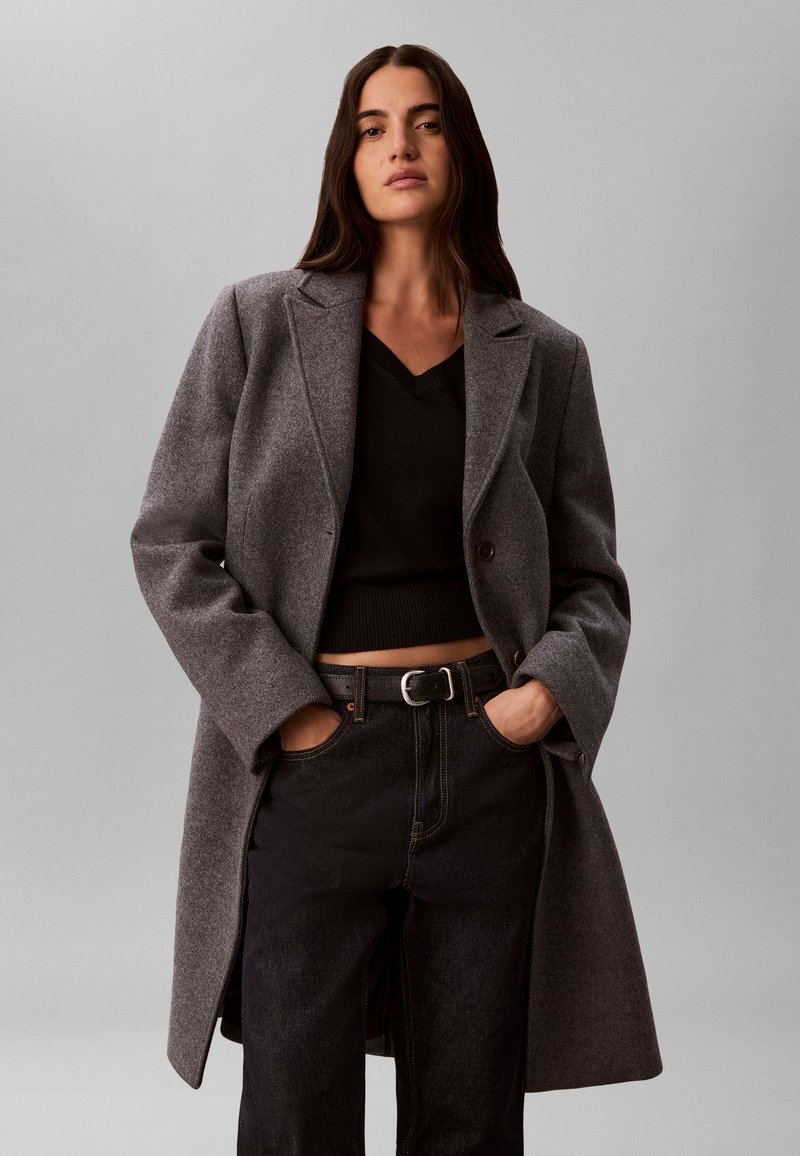 Femme aux longs cheveux foncés porte un manteau en laine gris, un pull court noir et un jean noir, les mains dans les poches, sur un fond gris uni.