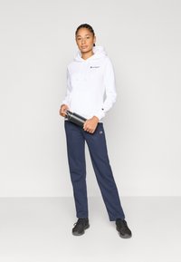 Λευκό φούτερ με κουκούλα, τσέπη καγκουρό και κορδόνια, συνδυασμένο με μπλε-σκούρο παντελόνι jogger. Κρατάει ένα παγούρι από ανοξείδωτο ατσάλι. Μαύρα αθλητικά παπούτσια.