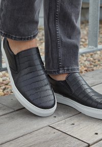 Baskets noires à enfiler avec motif texturé portées avec un jean gris sur une terrasse en bois près de gravier et d'une rambarde en métal.
