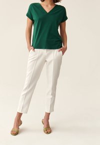 Vihreä v-kaula-aukkoinen t-paita, jossa on käännetyt hihat, yhdistetty valkoisiin muotoiltuihin housuihin, joissa on kapea leikkaus ja etuvälikkeet. Yhdistelmänä vihreät nilkkaketjulla varustetut korot.