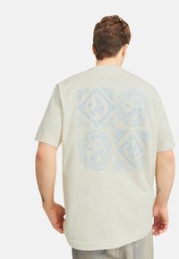 Kortærmet beige T-shirt med et design, der har blå cirkulære og diamantmønstre på ryggen, lavet af bomuldsstof.