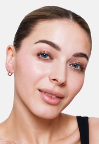 Goldene Ohrhänger mit strukturiertem Finish und klaren Strasssteinen. Das Modell hat helle Haut, glattes Haar und natürliches Make-up vor einem weißen Hintergrund.