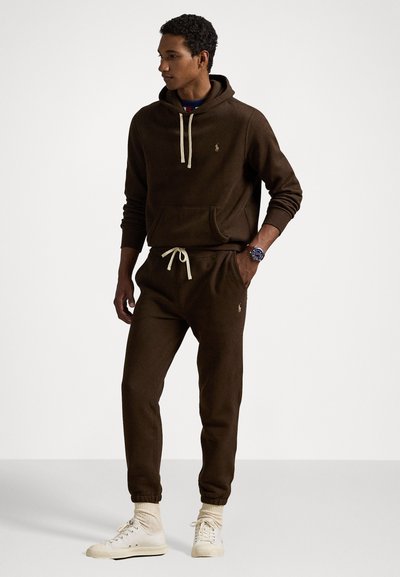 Brun hettegenser med beige snorer og logo, kombinert med matchende joggebukser. Antrekket har en avslappet passform og ribbestrikkede mansjetter.