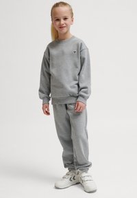 Grå sweatshirt och träningsbyxor set, tillverkat av mjuk bomull. Lätt lös passform, ribbade muddar och en liten svart logotyp på bröstet. Vita sneakers.