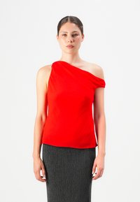 Mujer con una blusa roja brillante de un solo hombro y una falda negra texturizada, de pie frente a un fondo blanco liso.