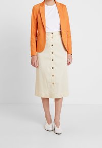 Oranssi bleiseri valkoisen t-paidan päällä, yhdistettynä vaaleanbeigeen hameeseen, jossa on edessä napit ja sileä rakenne. Valkoiset kengät täydentävät asua.