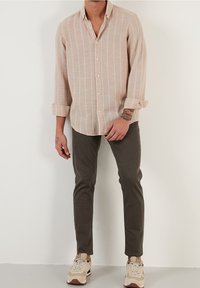 Světle béžová košile na knoflíky s dlouhým rukávem a bílými svislými pruhy, doplněná tmavě šedými kalhotami a béžovými teniskami. Ležérní, přiléhavý styl.