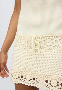 Κρεμ πλεκτό φόρεμα με ριμπ ύφανση και δαντελένιο τελείωμα κροσέ· διαθέτει λουλουδένια σχέδια και κ ορδόνι στη μέση για ρυθμιζόμενη εφαρμογή.