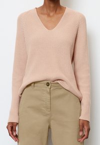 Pull en maille rose clair avec un col en V, une texture côtelée et des manches longues. Associé à un pantalon beige, avec une fermeture à boutons.