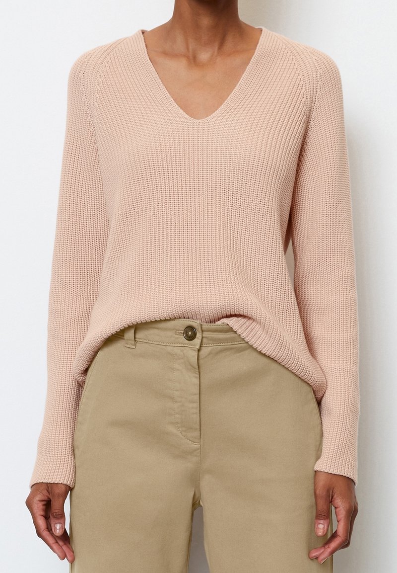Pull en maille rose clair avec un col en V, une texture côtelée et des manches longues. Associé à un pantalon beige, avec une fermeture à boutons.
