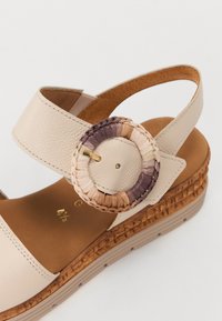 Beige leather sandal with a wide strap, textured insole, and a circular woven buckle featuring colors of cream, pink, and purple accents.