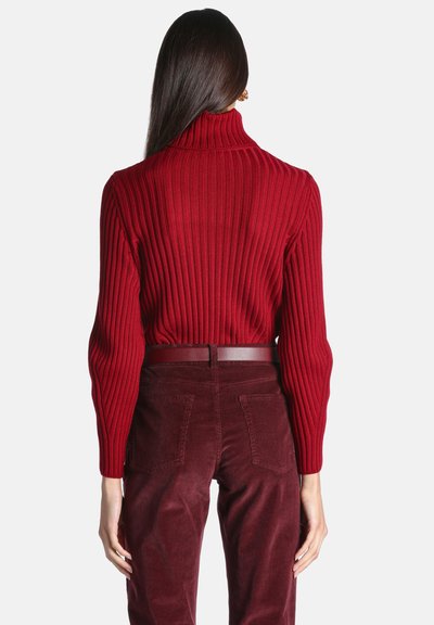 Femme aux longs cheveux foncés porte un pull à col roulé rouge côtelé rentré dans un pantalon en velours côtelé rouge foncé taille haute avec une ceinture assortie.