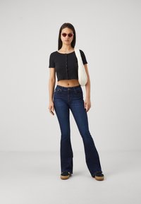 Black cropped t-shirt with buttons, dark blue flared jeans, brown platform shoes, and light-colored shoulder bag. Pink sunglasses.