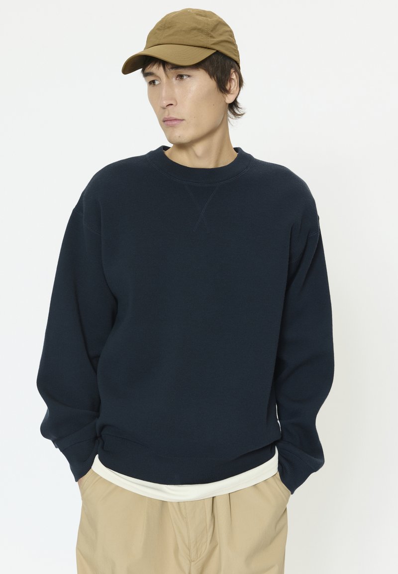 Sweat-shirt bleu marine à col ras du cou et poignets côtelés, assorti d'un t-shirt de couleur claire et d'un pantalon kaki. Porté avec une casquette beige.