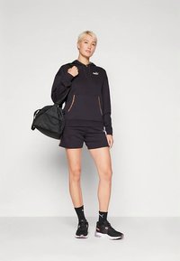 Czarna bluza z kapturem z logo, czarne spodenki, czarne sneakersy oraz czarna torba. Zauważalne kontrastowe przeszycia i kieszenie. Codzienny strój sportowy.