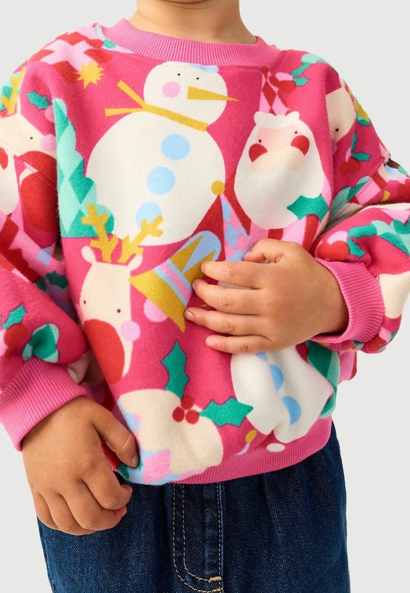 Enfant portant un pull rose avec des motifs de Noël colorés comprenant un bonhomme de neige, le Père Noël, un renne, du houx et une cloche, assorti à un jean bleu.