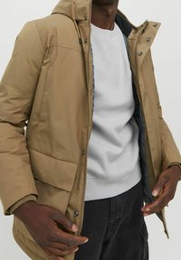 Beige isolerad jacka med huva, framdragkedja och tryckknappsfickor. Har en grå foder och ribbade ärmslut. Bärs över en grå sweatshirt.