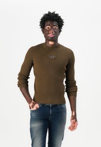 Olivgrüner, gerippter Pullover mit langen Ärmeln, Rundhalsausschnitt und kleinem Logopatch. Slim Fit und strukturiertes Strickmuster. Kombiniert mit blauen Jeans.
