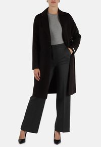 Cappotto lungo nero su un top grigio, abbinato a pantaloni grigi su misura e tacchi neri. Tessuto morbido con linee pulite e una vestibilità strutturata.