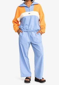 Felpa con zip color block in arancione, bianco e azzurro chiaro; abbinata a pantaloni a gamba larga azzurri chiari; tessuto morbido, vestibilità rilassata.