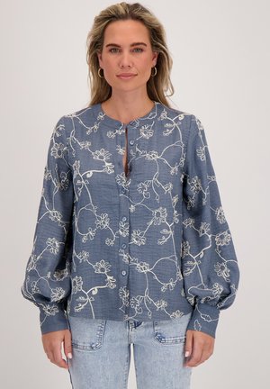 Vrouw in een blauwe blouse met wit bloemenborduursel en opgezette mouwen, gecombineerd met licht gewassen jeans voor een effen achtergrond.