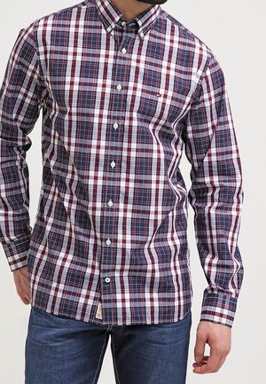 Homme portant une chemise à manches longues à carreaux boutonnée en bleu marine, blanc et rouge, associée à un jean bleu, debout devant un fond uni.