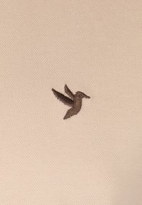 Broderie d'un colibri marron sur un tissu texturé beige clair, avec des points lisses et réguliers et un design simple et minimaliste.