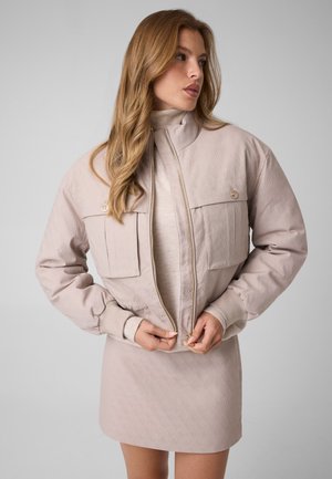 Un mannequin portant une veste beige clair et une jupe assortie ajuste la fermeture éclair de la veste devant un fond de studio gris uni.