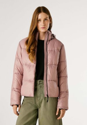 Doudoune rose clair avec fermeture éclair à l'avant, capuche et quilting horizontal. Portée par-dessus un haut noir et un pantalon vert olive.